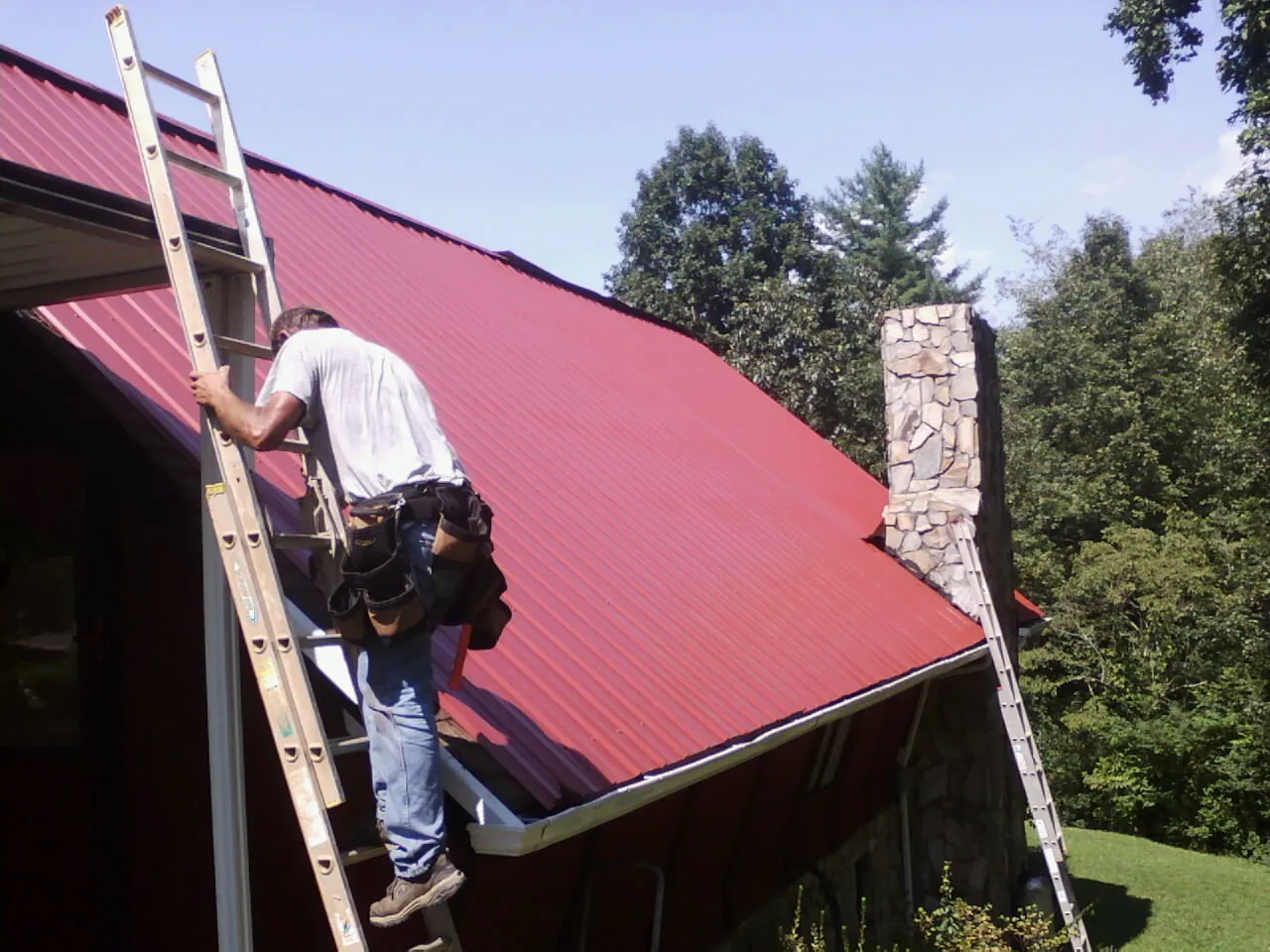 Roofer on a ladder beside a red metal roof and stone chimney — Soffit & Fascia Repair by Roofing Brothers in American Falls
